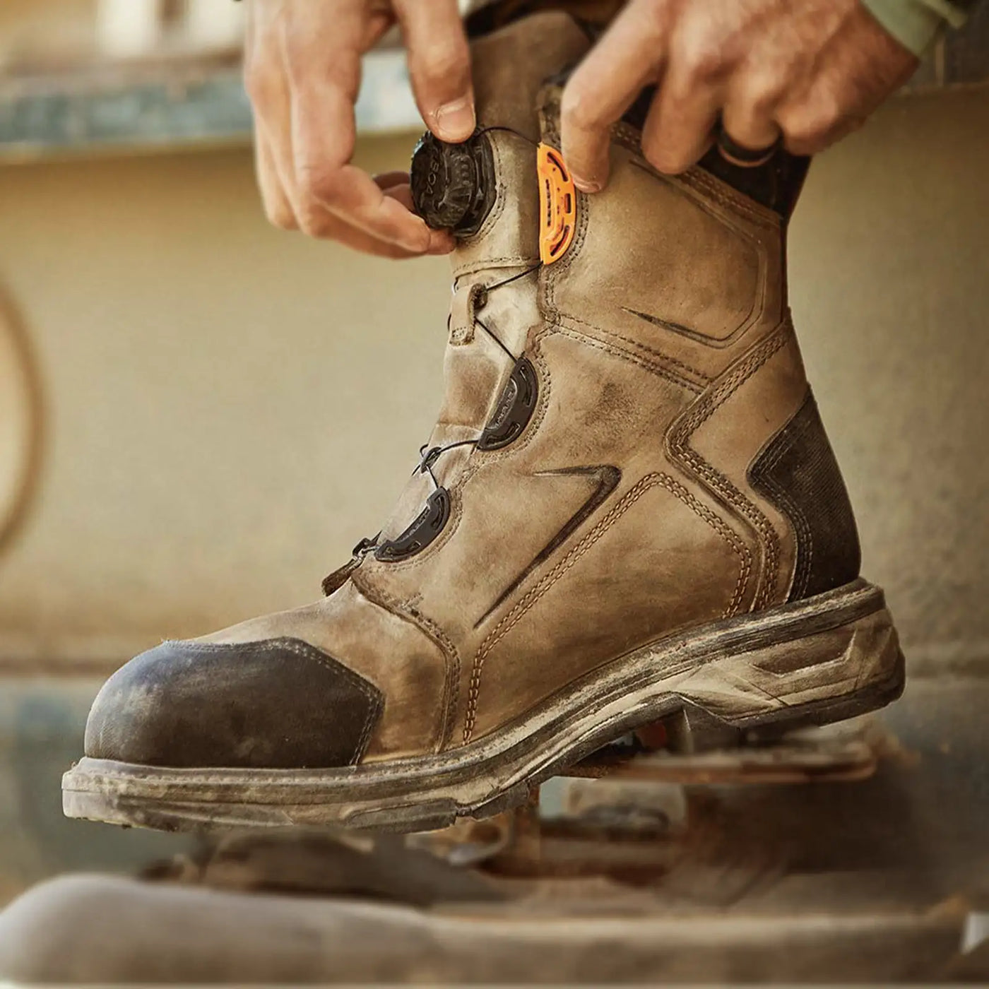 Hands tightening a dusty BOA® work boot with dial closure on a jobsite.