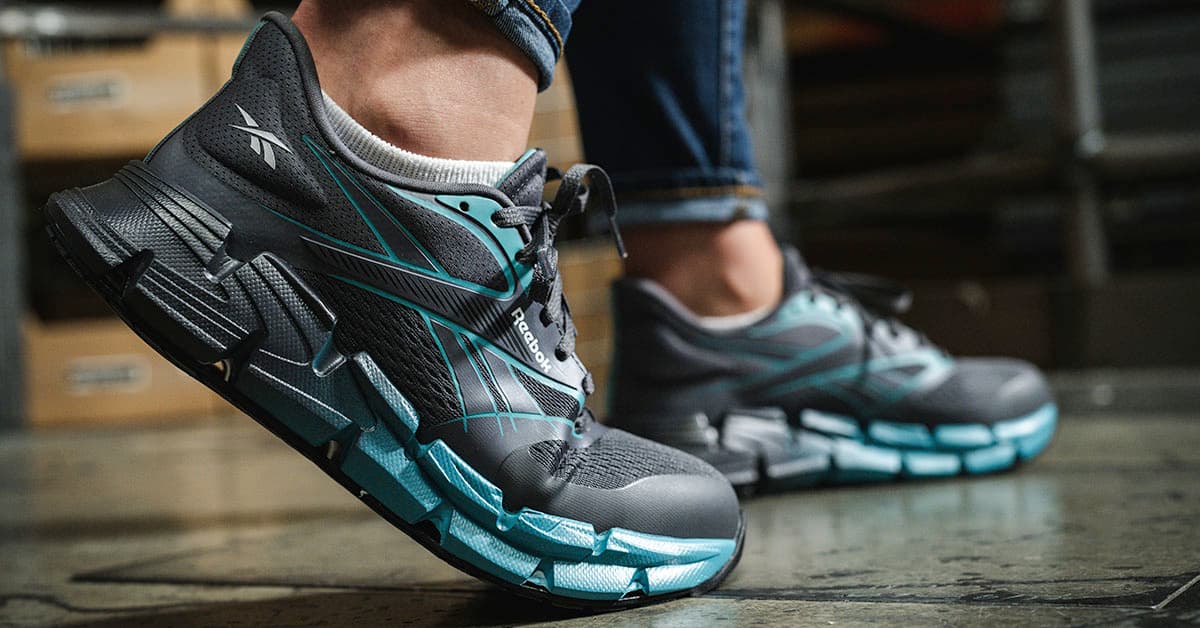 Reebok athletic work shoes and boots displayed on a warehouse floor with the Reebok logo overhead.
