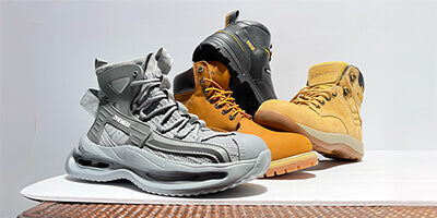 Three pairs of safety work boots in beige, black, and brown, displayed outdoors on a stone surface with greenery and a metal fence in the background