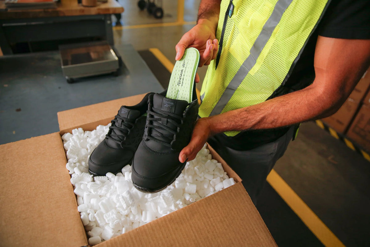 Worker holding black Reebok work shoes and a removable MemoryTech Massage insole over a shipping box